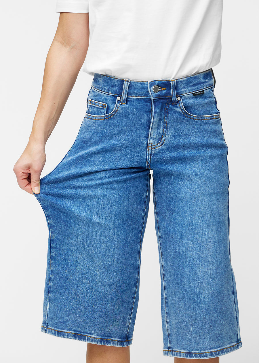 Model strækker de løse, klassisk blå denim capris i farven Rivers ved hoften for at vise materialets stretch.