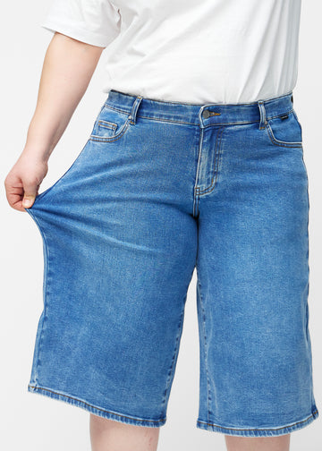 Plus-size model strækker de løse, klassisk blå denim capris i farven Rivers ved hoften for at vise materialets stretch.