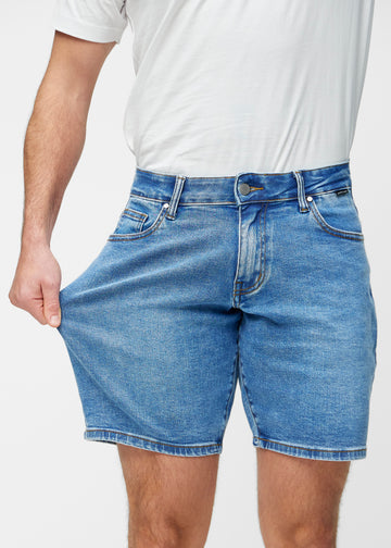 Model stretches the blue denim shorts for men in the colour Rivers at the thigh to show the stretch of the material.