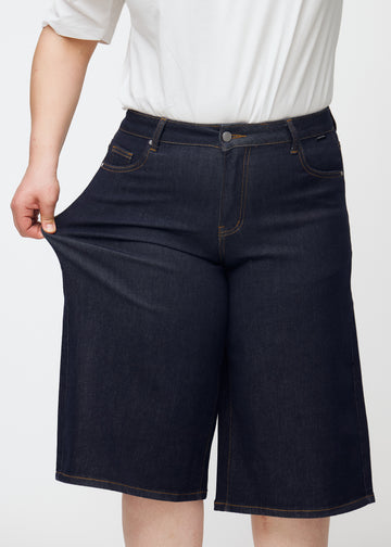 Plus-size model stretches deep dark blue denim capris for women in the colour Midnights at the thigh to show the material’s stretch.