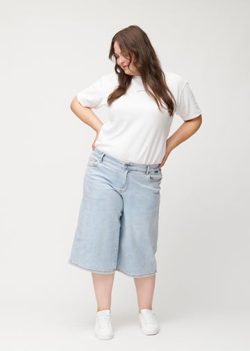 Fuldt billede af plus-size model i løse, lyseblå denim capris i farven Waves, der går til lige under knæet.