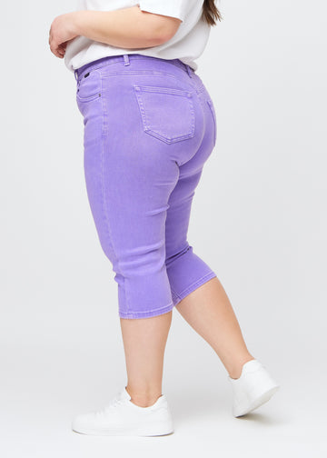 Slim capris for women in pastel purple jeans fabric, color Lavenders. Side view on a plus-size model, showing the tailored fit and mid-rise waist.