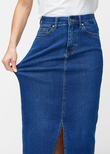Model strækker den lange, mørkeblå denimnederdel i farven Oceans ved hoften for at vise materialets stretch.