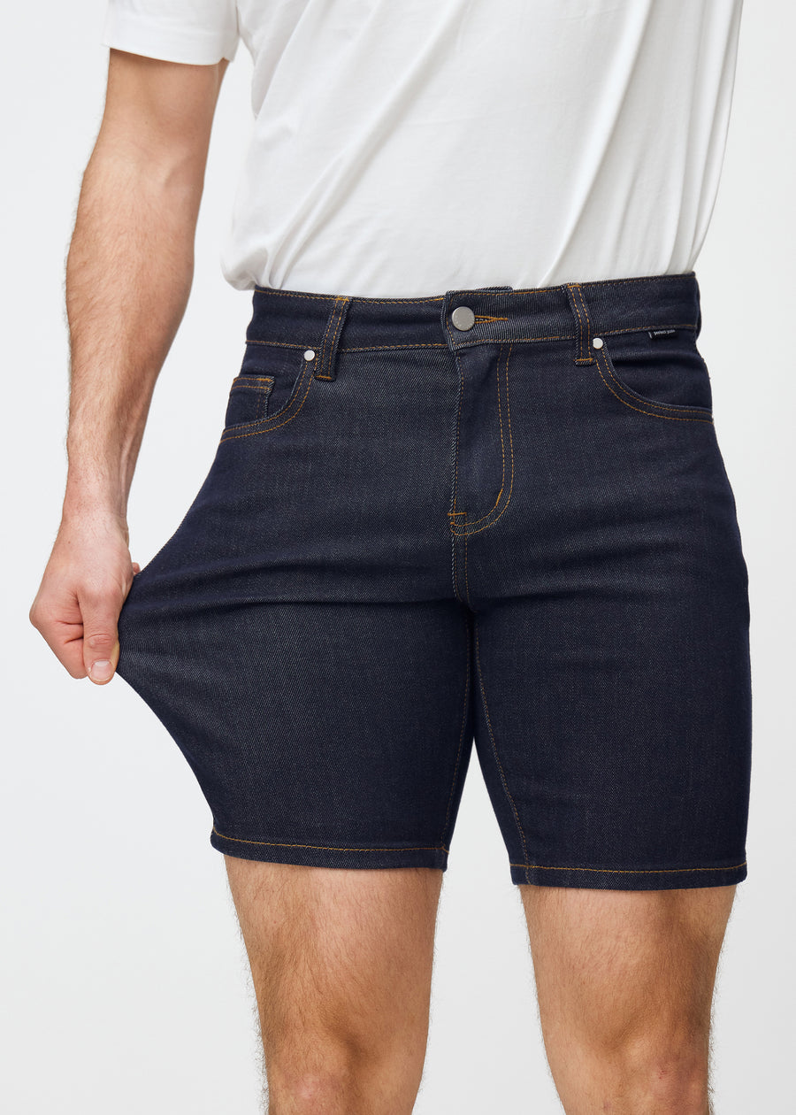 Model stretches deep dark blue denim shorts for men in the colour Midnights at the thigh to show the material’s stretch.